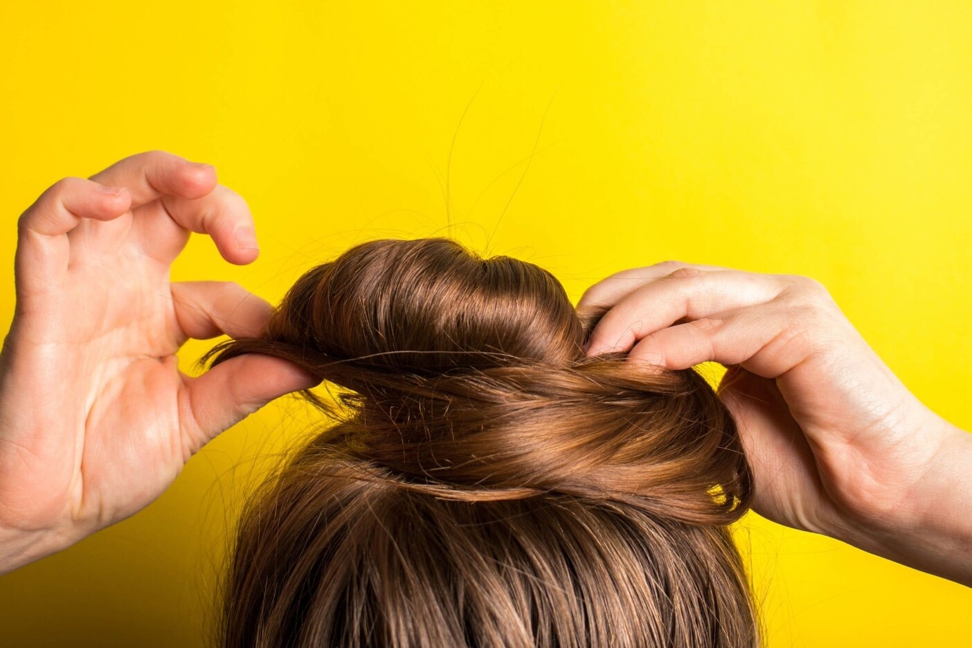 3-easy-summer-hairstyles-for-hot-days 2400 x 1600_edited Close-up of hands arranging brown hair into a bun against a bright yellow background. The person is in the process of styling their hair, with fingers holding and shaping the bun, perhaps inspired by a visit to one of the best hair salons Cary, NC has to offer.