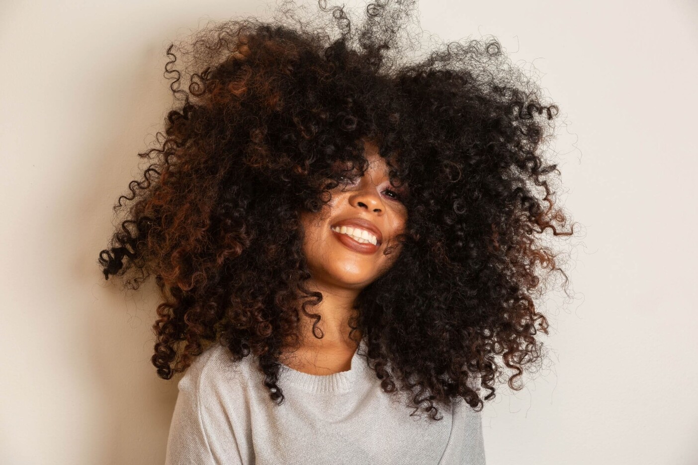 hairstyle-trends-for-2021 2400 x 1600_edited Smiling person with voluminous, curly hair that obscures part of their face, likely freshly styled at a hair salon. They are wearing a light grey top and are positioned against a plain white background.