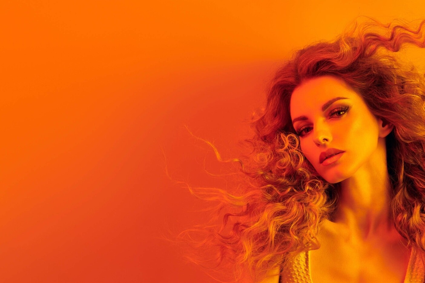 A person with voluminous, curly hair poses against a vibrant, orange gradient background. They have an intense expression and are looking slightly off to the side. The overall image is bathed in warm, orange tones, creating a dramatic and bold effect, reminiscent of trendy styles found at top hair salons near me.