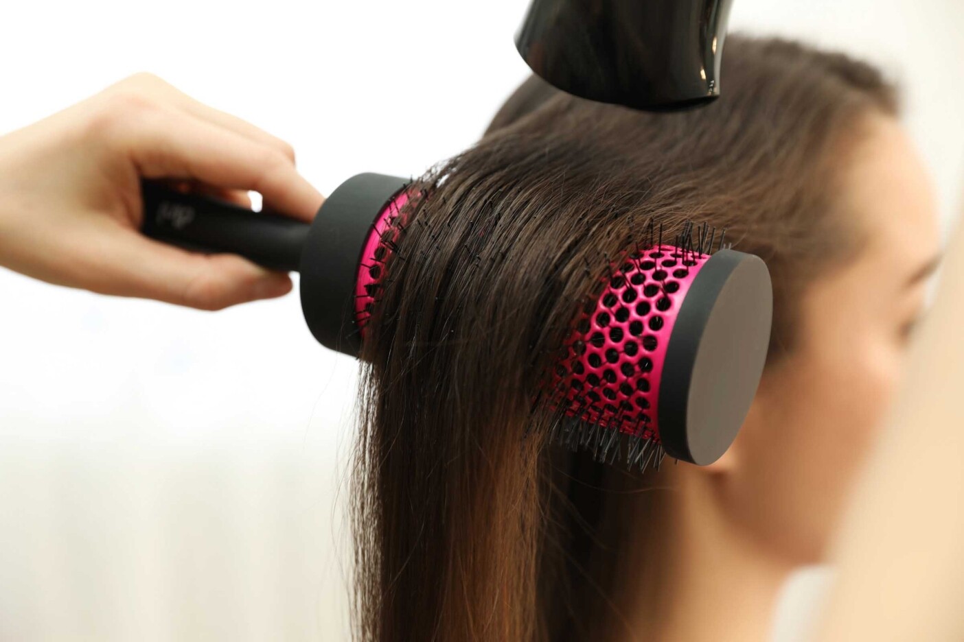 Close-up of a person having their hair styled at one of the best hair salons Cary NC offers. A hand holds a round hairbrush with a pink and black handle, while a hairdryer is used to dry the long, straight hair, brushing and styling it simultaneously.