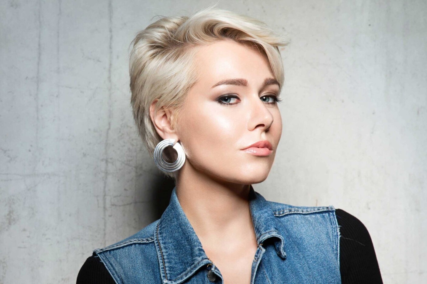 A person with short, styled blonde hair, fresh from a Sunday salon session, wearing bold silver hoop earrings and a denim vest over a dark top, poses confidently against a plain, textured background. Their expression is serious and self-assured.