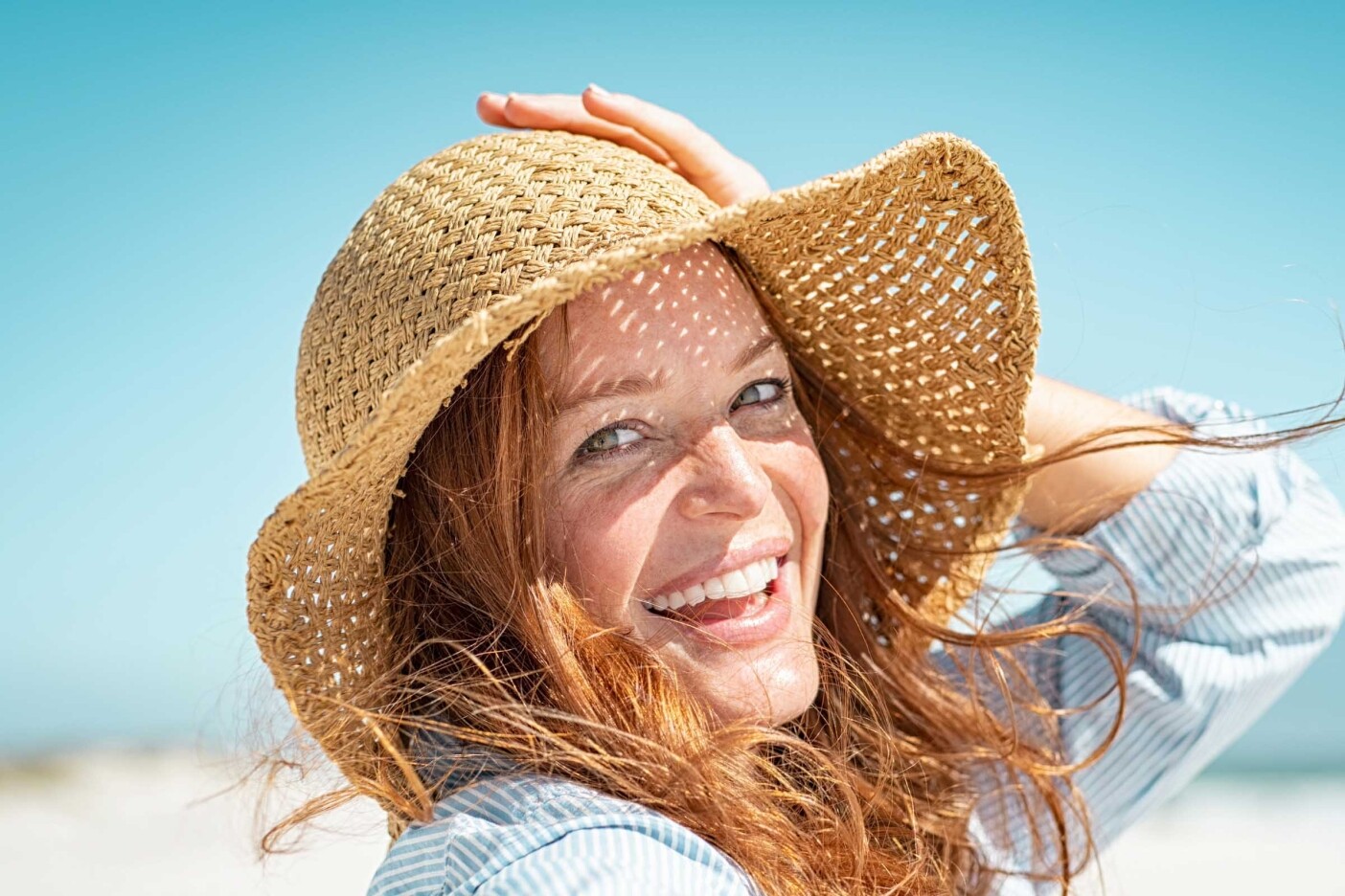 6 Expert Tips from Cary’s Top Hair Stylists for Gorgeous Summer Hair Blog Post Header 2400 x 1600_edited A woman with long, summer hair, wearing a straw hat, smiles brightly under a clear blue sky. The beach is blurred in the background, suggesting a sunny day. She holds the brim of her hat with one hand as her hair flows gently in the breeze—hair stylists' tips brought to life.