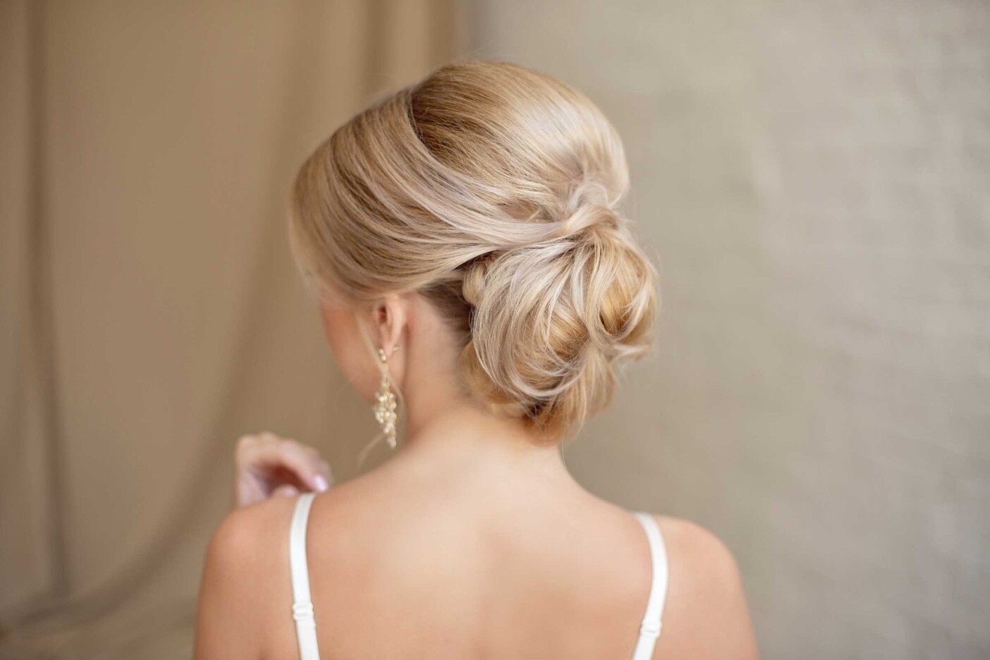 Elevate Your Event Look with Tips from a Professional Hair Stylist Blog Post Header 2400 x 1600_edited A woman with blonde hair styled in an elegant updo, featuring soft waves and a loose bun, is shown from behind. She is wearing a white spaghetti strap top and gold dangling earrings. The background is softly blurred with neutral tones, showcasing the expertise of formal hair styling services in Cary, NC.