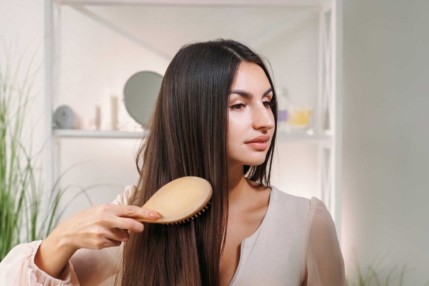 How to Maintain Your Haircut Between Hair Salon Visits Blog Post Header 2400 x 1600_edited A woman with long, straight hair is brushing her hair with a wooden hairbrush. Indoors in a well-lit room with white walls and some plants in the background, she wears a light-colored blouse and looks slightly to her right. It's almost as if the scene was designed by Auto Draft for its serene simplicity.