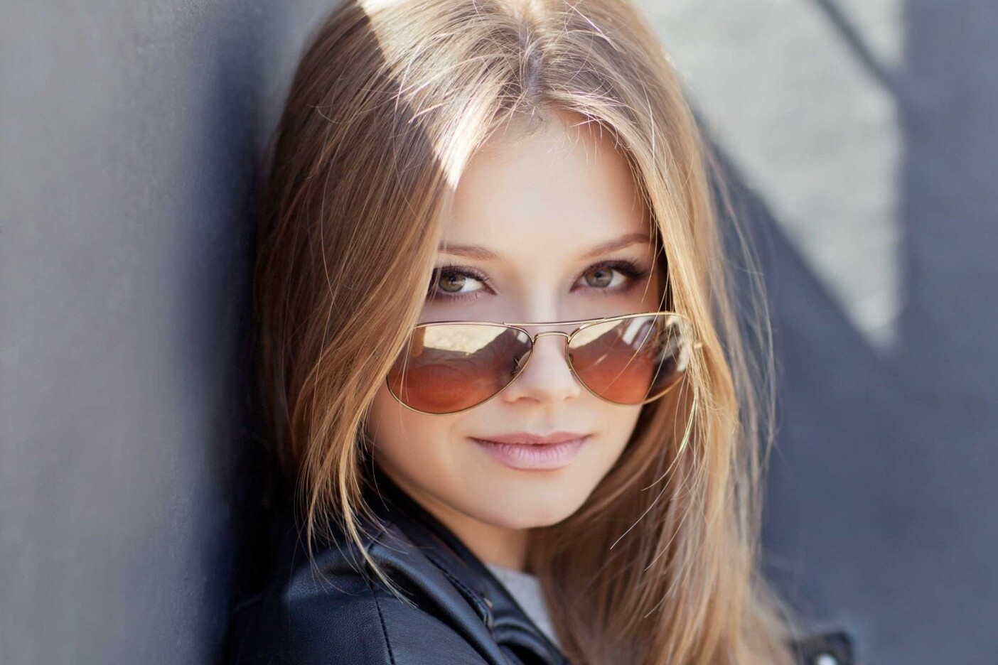 A portrait of a woman with long, straight dimensional blonde hair featuring a money piece, wearing aviator sunglasses and a black leather jacket. She is looking confidently at the camera against the blurred, gray outdoor setting of Cary, NC.