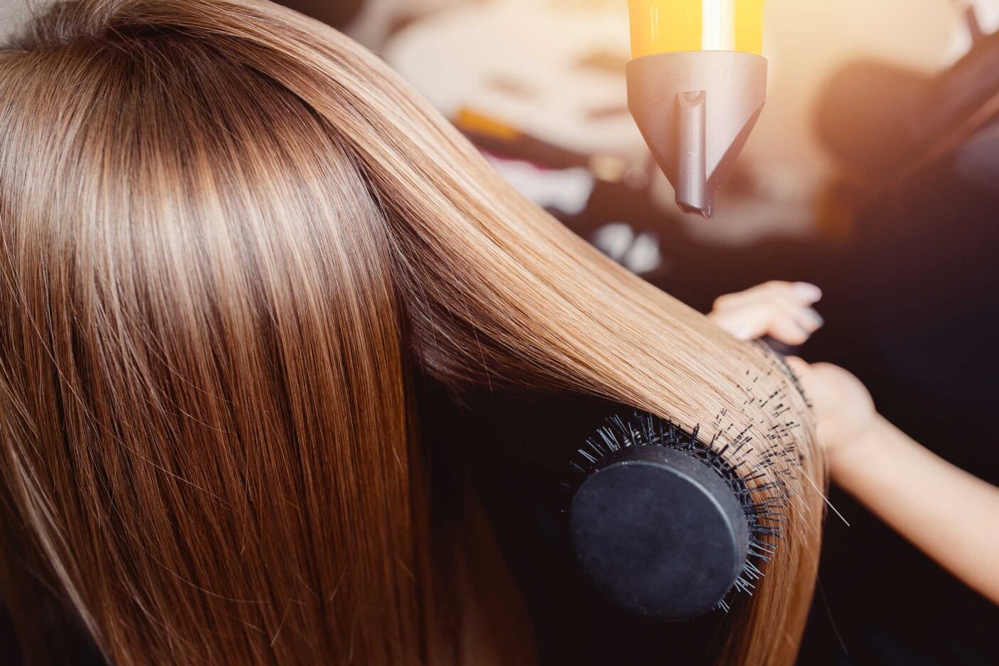 A person with long, straight, blonde hair is having their hair blow-dried during their first visit to the hair salon. A round brush is being used to style the hair while a hairdryer is directed at the brush. The scene appears to be in a salon or a home setting.