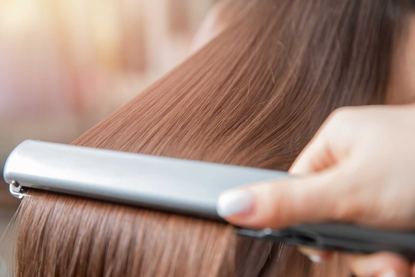 A close-up of a person using a flat iron to straighten long, light brown hair treated with keratin. The hand holding the flat iron has neatly manicured nails painted a light color. The hair appears smooth and shiny, reflecting light gently.