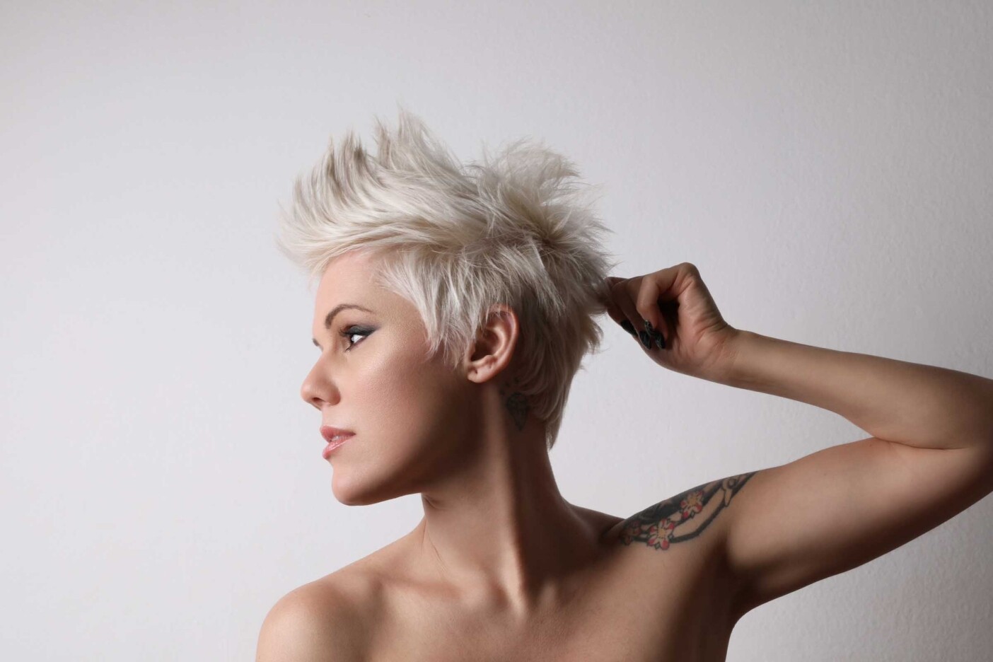 why-a-professional-haircut-is-worth-the-investment Blog Post Header 2400 x 1600_edited A person with a professional haircut—a short, platinum blonde mohawk—is looking to the side, touching their hair with one hand. The individual has a visible tattoo on their upper arm. The background is plain white.