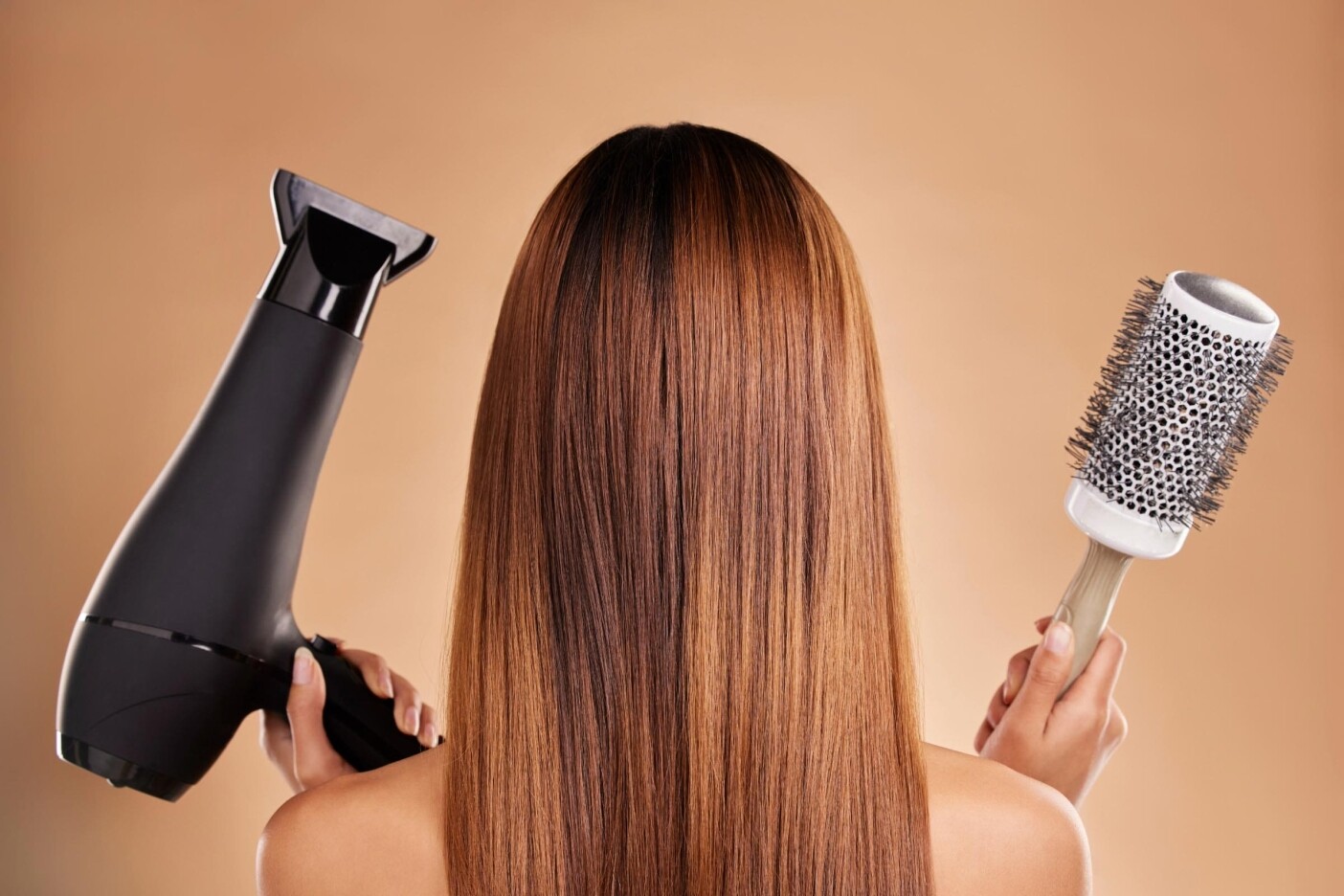 10 Hair Styling Tips for a Modern Look Blog Post Header 2400 x 1600_edited A person with long, straight brown hair holds a black hairdryer in one hand and a round hairbrush in the other, skillfully achieving a modern look against a warm brown background. Perfect for those seeking professional hair styling tips.