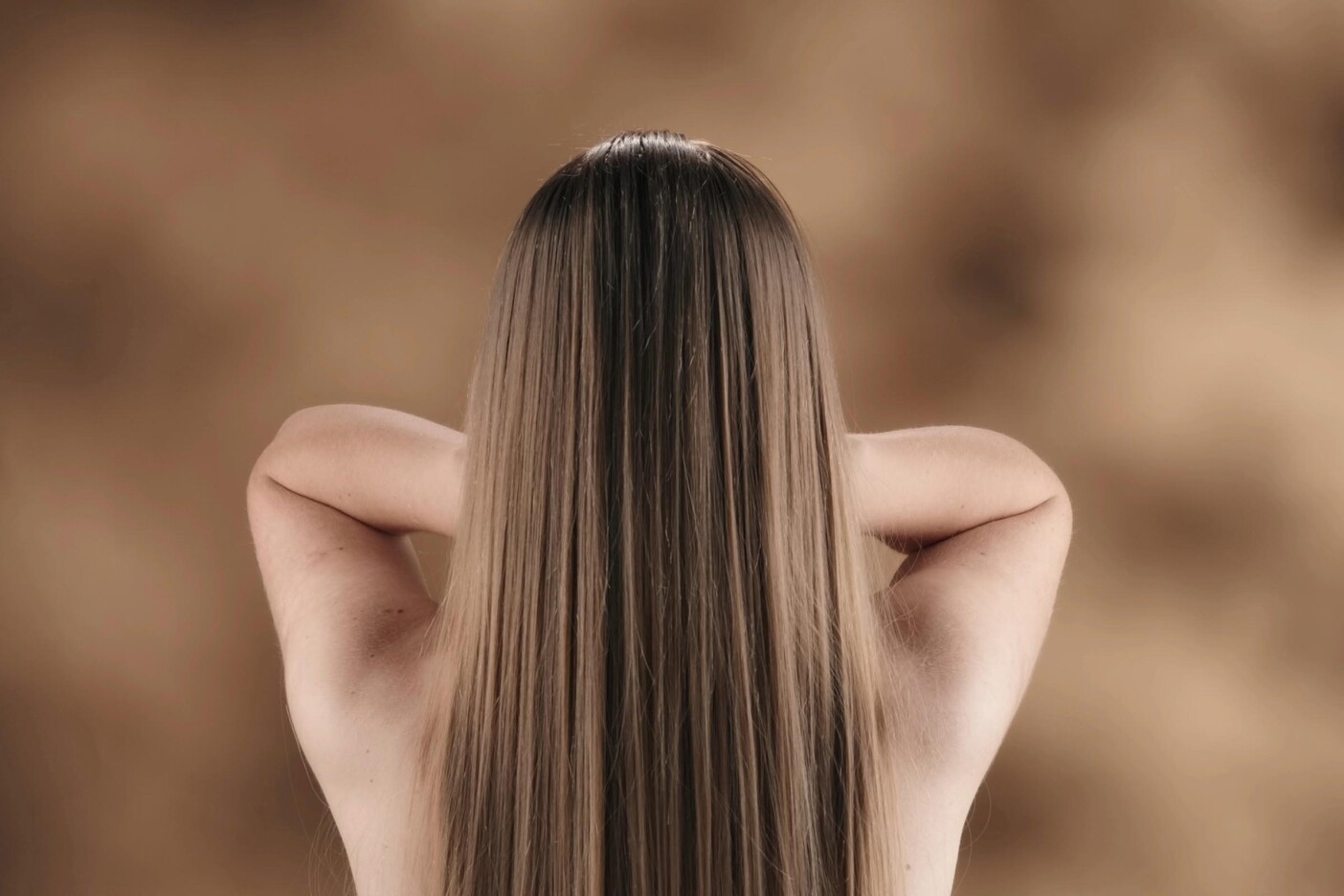 Why Dirty Blonde Hair is the Ultimate Versatile Shade Blog Post Header 2400 x 1600_edited A person with long, straight, dirty blonde hair stands with their back to the camera, arms raised and hands resting lightly on the back of their neck against a soft, brown blurred background.