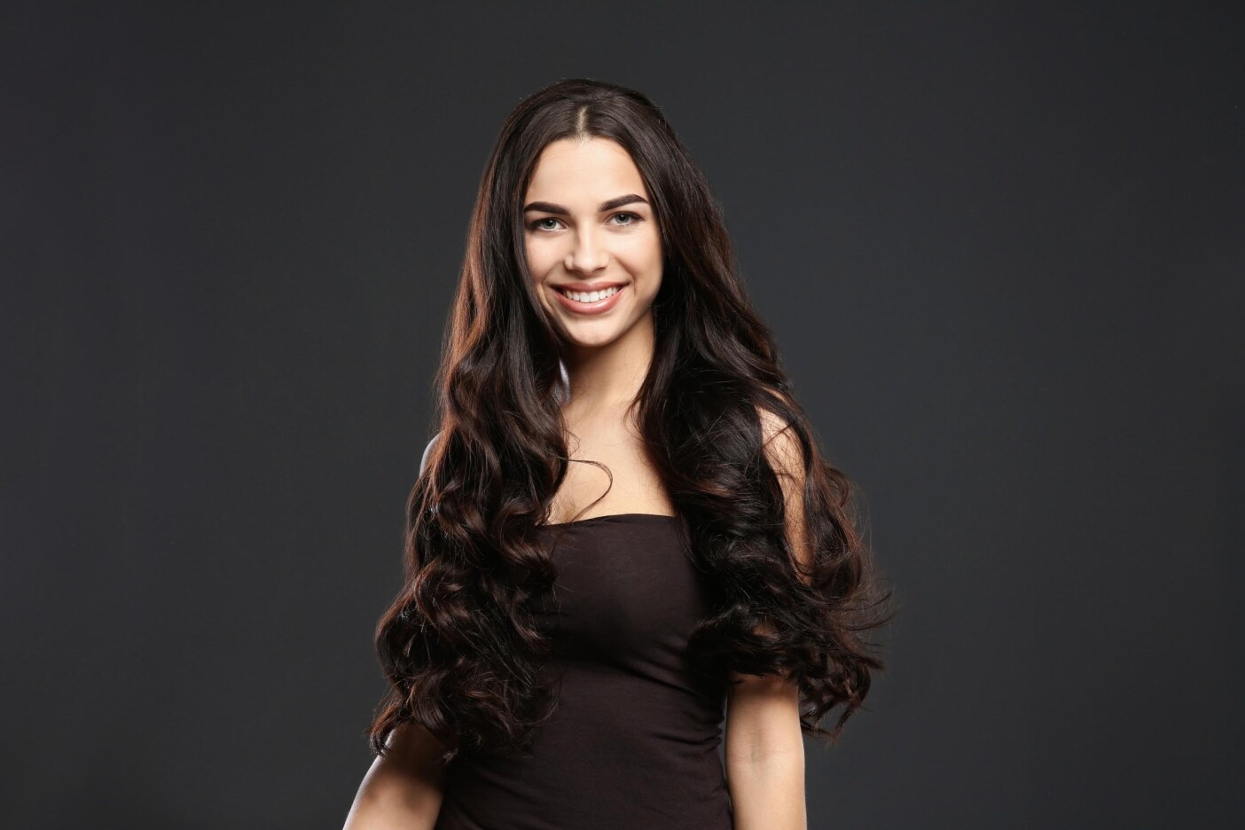how much are hair extensions 2400 x 1600_edited Smiling woman with long, wavy dark hair wears a sleeveless black top, standing against a dark background.