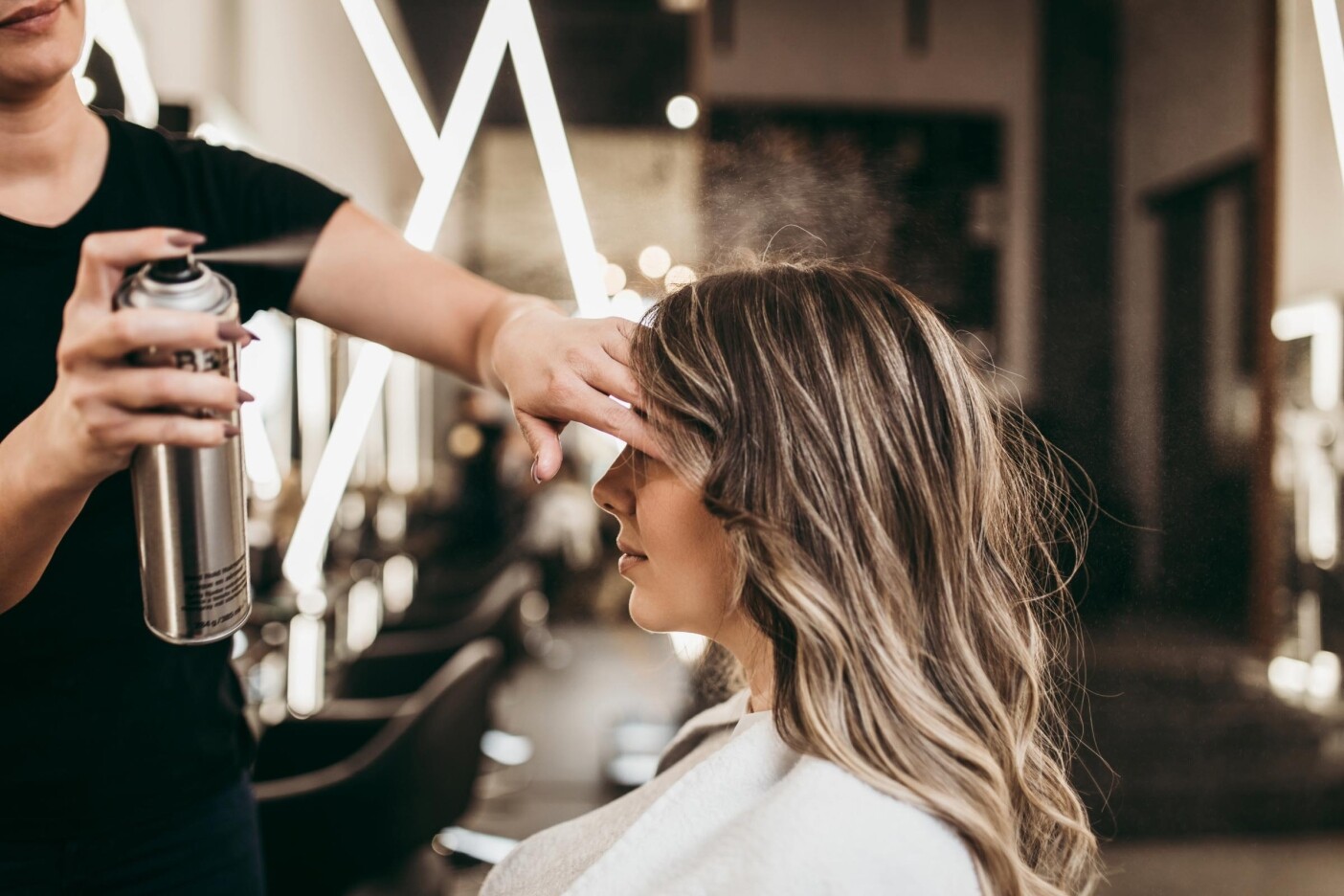 A woman with long, wavy hair sits in one of the best hair salons Cary, NC offers, as a stylist applies hair spray. Bright, geometric lights illuminate the scene, creating a modern atmosphere.
