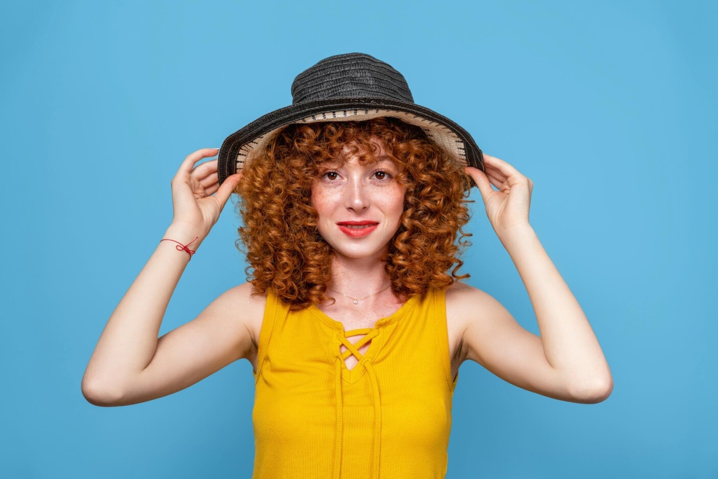 A person with curly red hair, reminiscent of wild perms, smiles while holding the brim of a wide-brimmed black hat. They are wearing a yellow sleeveless top and are set against a plain blue background.