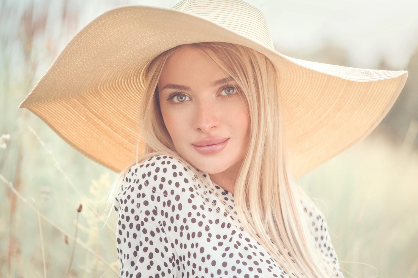 How Can I Maintain the Shine of My Blonde Hair at Home_edited A woman with long blonde hair is wearing a large, wide-brimmed straw hat and a white blouse with black polka dots. She is outdoors, surrounded by soft-focus greenery, giving the image a warm, serene feel.