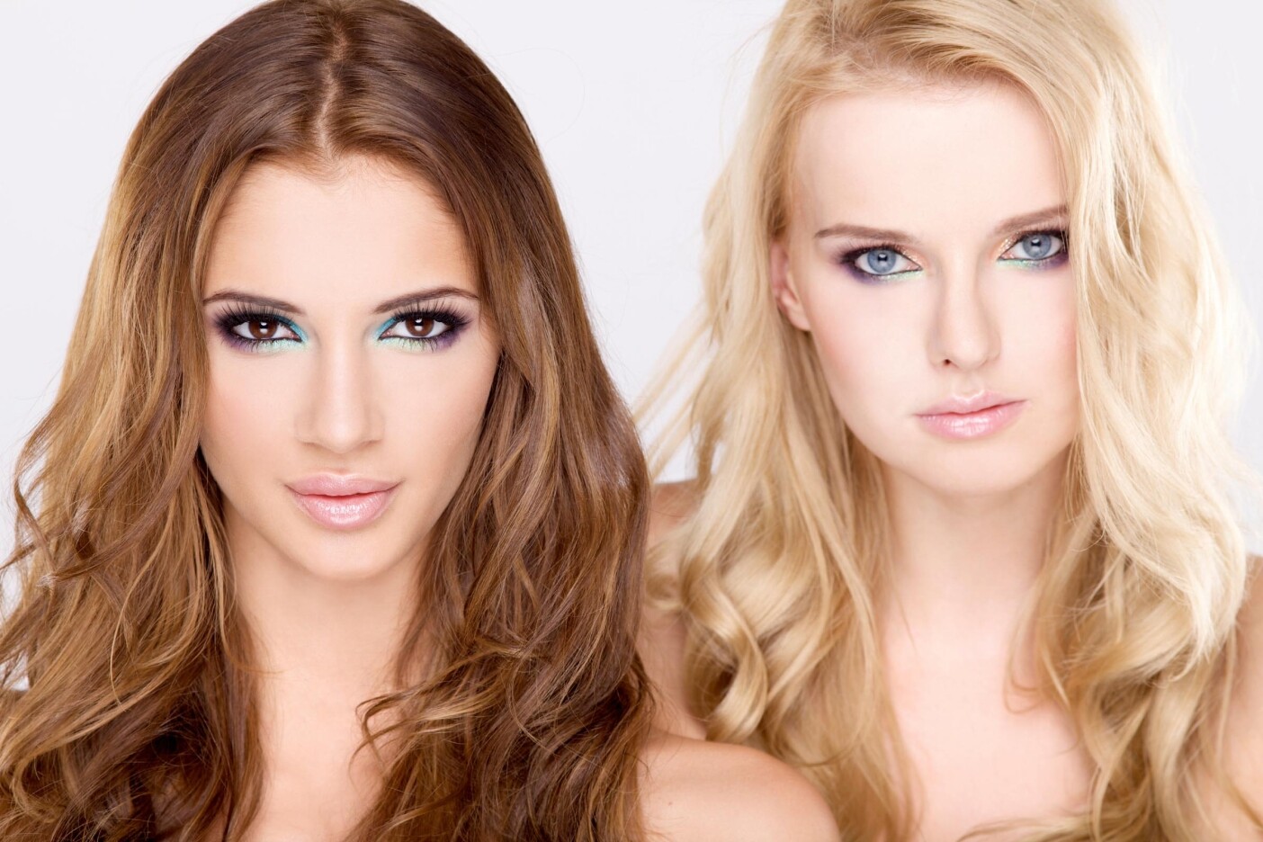 Two women with long, wavy hair showcase their distinct hair color. The brunette on the left has brown tresses, while the blonde on the right flaunts golden locks. Both sport bright makeup focusing on their eyes, maintaining a neutral expression against a plain background.