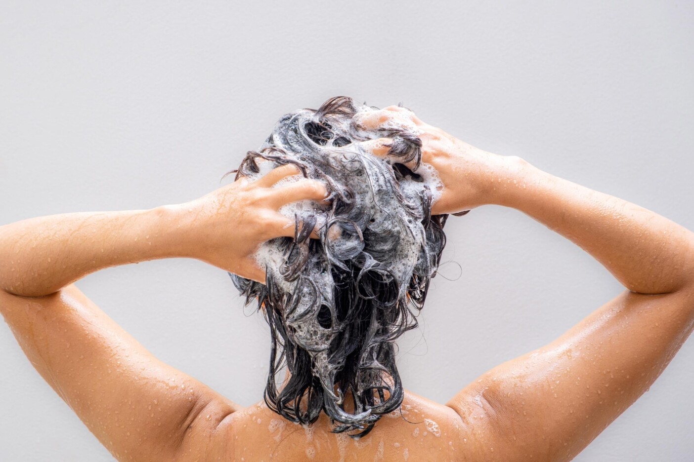 A person with wet hair seen from behind is washing their hair, creating a lather of shampoo with both hands—a great example of how to wash curly hair. Water droplets glisten on their arms and back against a plain light background.