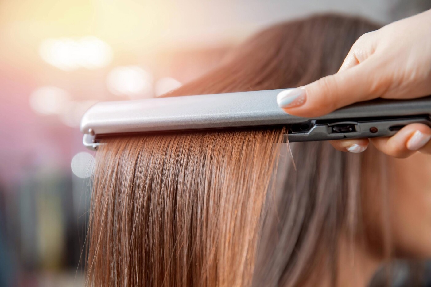 Close-up of a person straightening brown hair with a silver hair straightener, highlighting smooth, shiny strands and proper hair care after keratin treatments. A hand firmly holds the device for sleek results.
