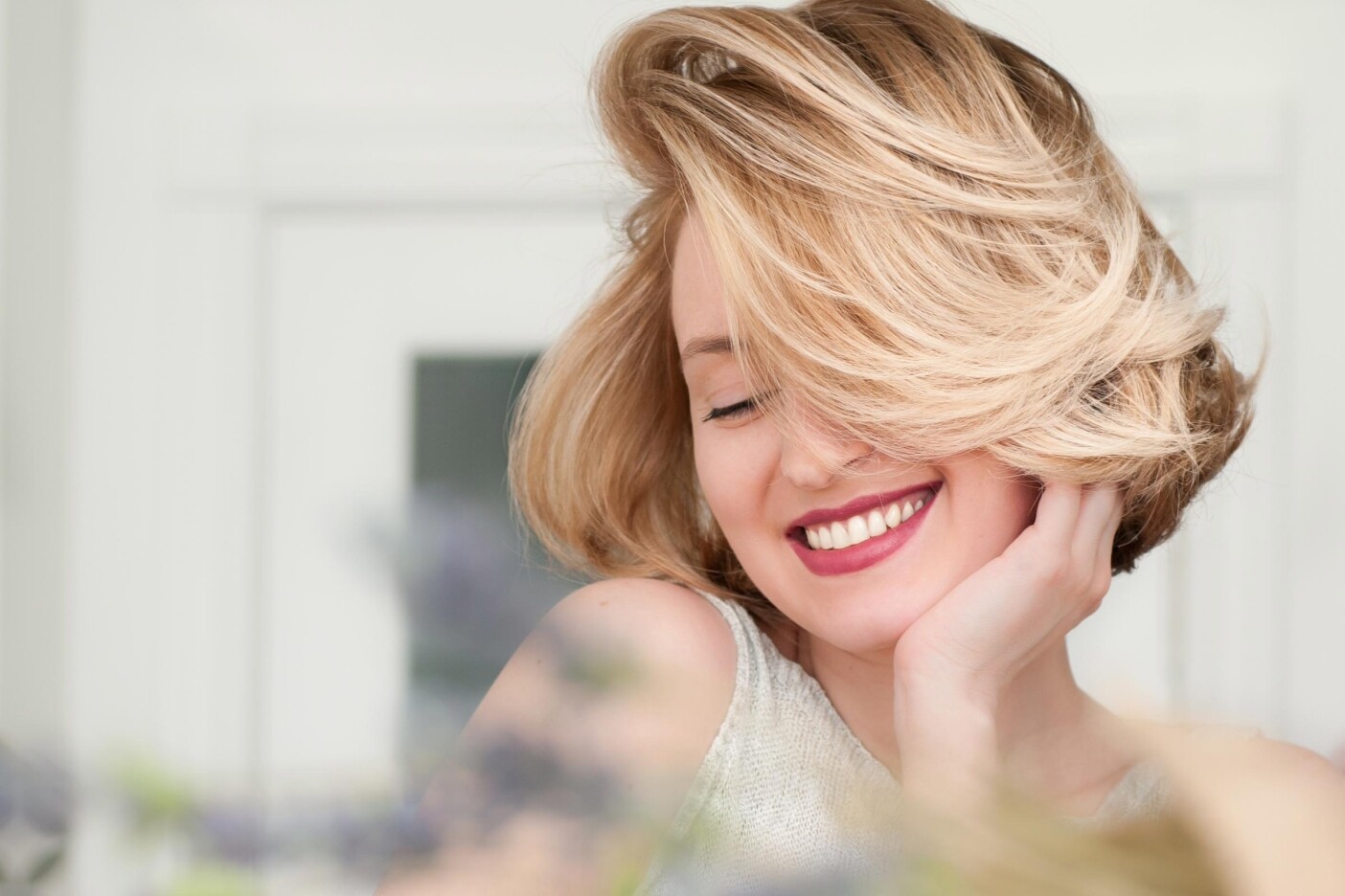 style.short.hair A woman with short hair and wavy blonde locks smiles with her eyes closed, resting her face in her hand. She wears a light-colored sleeveless top, and the background is softly blurred.