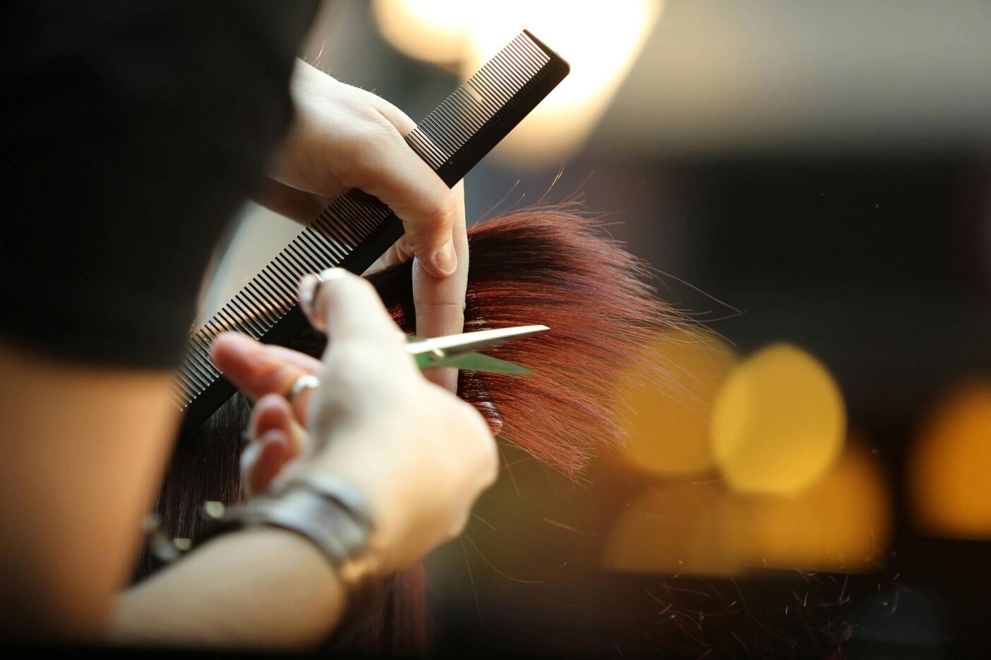 haircuts A close-up of a hairstylist’s hands cutting brown hair with scissors while holding a comb, with blurred lights in the background—perfect for those seeking haircut tips or inspiration to maintain a haircut at home.