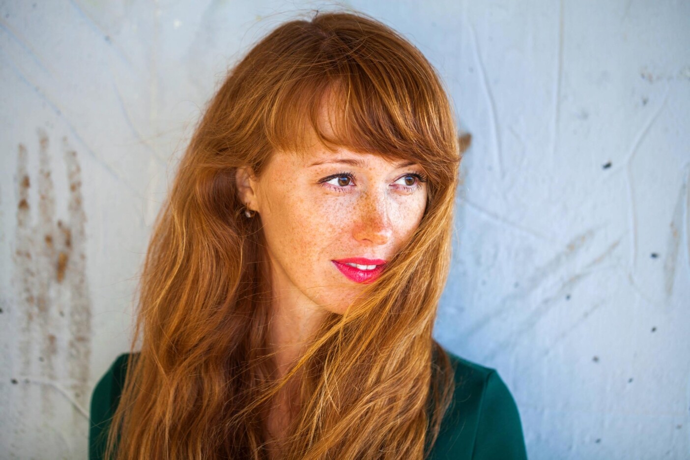 A woman with long red hair and freckles looks to the side, her hair partially covering her face. She is wearing a dark green top and bright red lipstick, standing in front of a light-colored textured wall.
