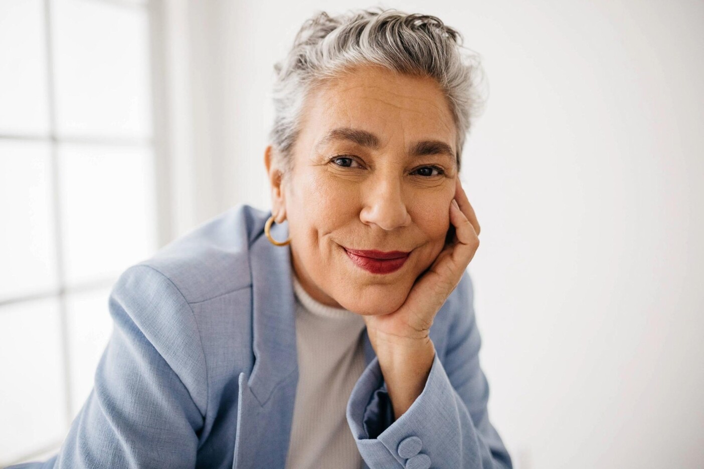 grey-7 A smiling older woman with short gray hair showcases stylish grey coverage as she rests her chin on her hand. She wears a light blue blazer, white top, gold hoop earrings, and red lipstick, with a bright window in the background.
