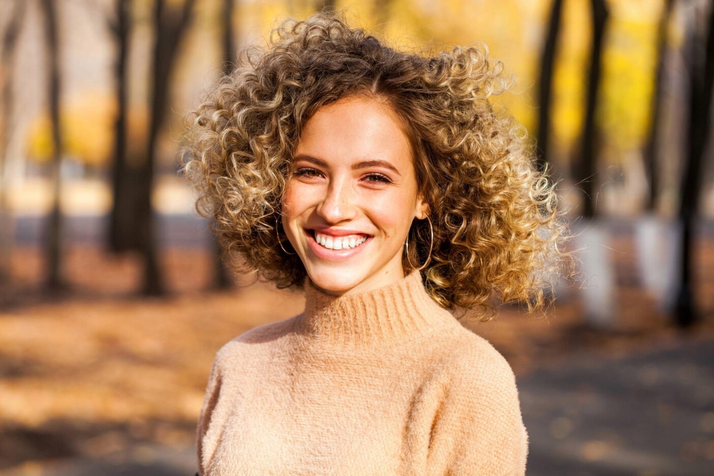 hair styling A young woman with curly blonde hair and a beige sweater smiles outdoors in a park, her hair styling perfectly framed by autumn leaves and sunlight that highlights her face and hair.