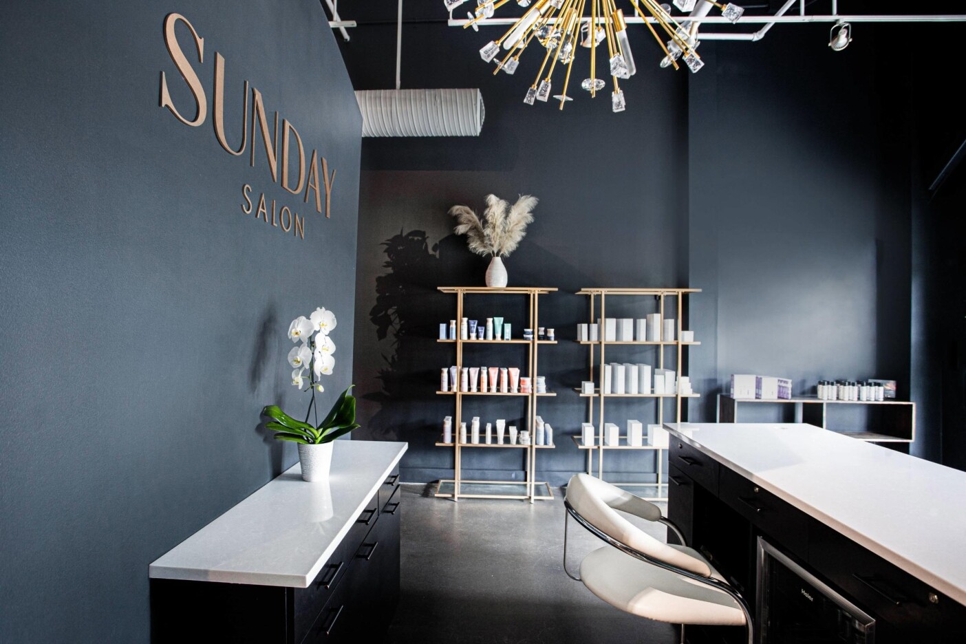 Modern salon interior with dark walls, a white counter, a white salon chair, and an orchid plant signals quality salon services. Beauty products line the shelves, pampas grass decorates the space, and a "SUNDAY SALON" sign shines overhead.