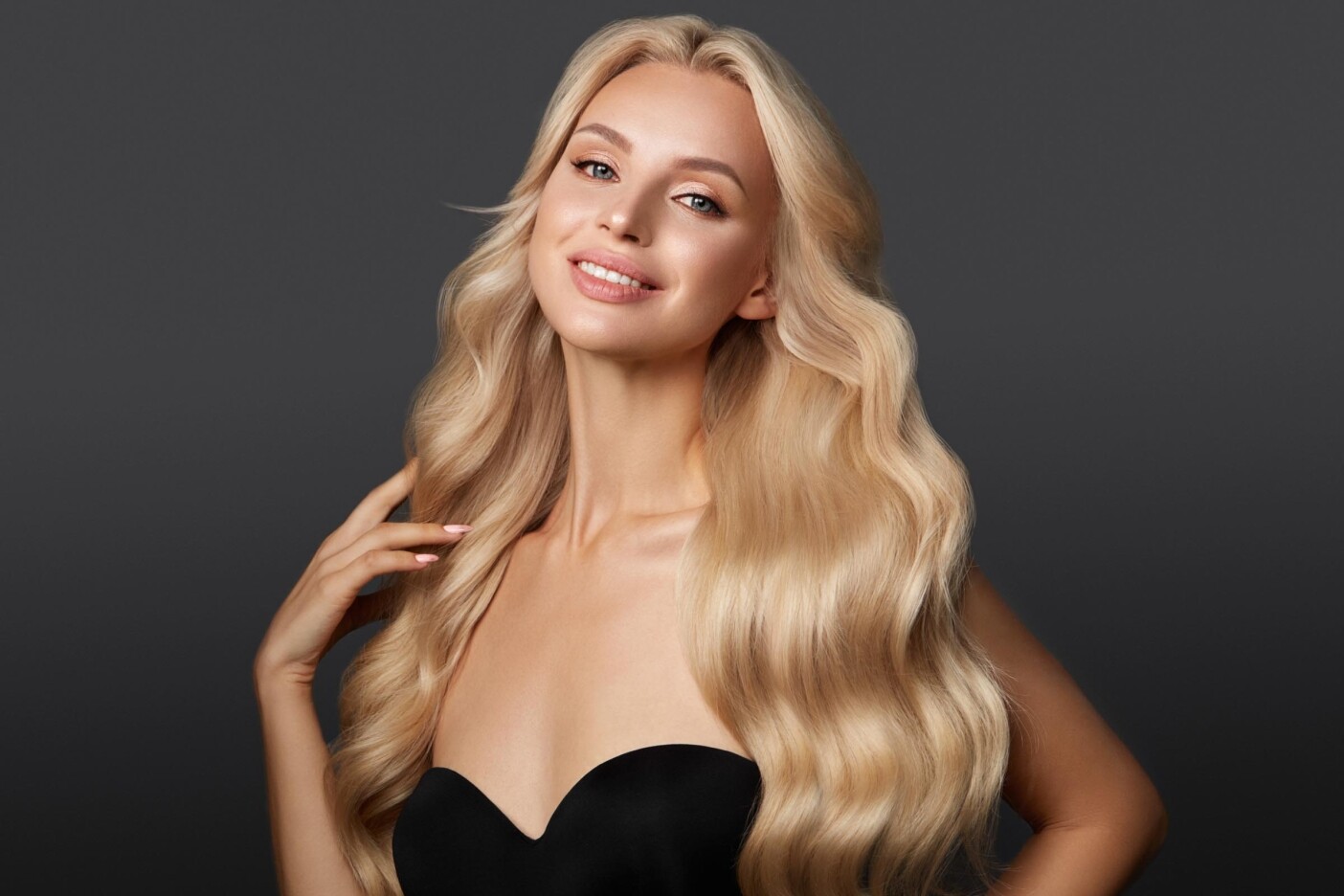 A smiling woman with long, wavy blonde hair—enhanced by flawless hair extension slide ups—poses against a dark background, wearing a strapless black top and looking confidently at the camera.