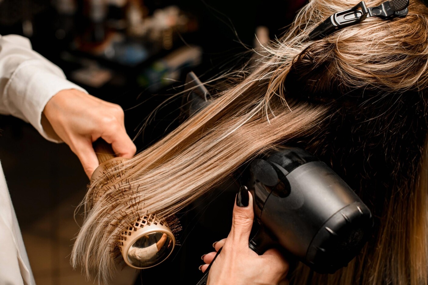 In a luxury hair salon, a stylist section-clips long blonde hair and expertly blow-dries it with a round brush and sleek black hairdryer for a flawless finish.