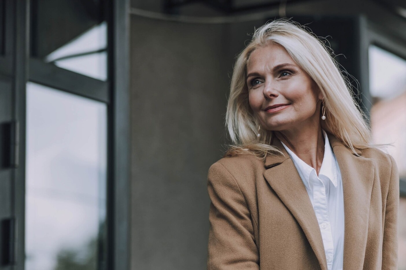 how-to-transition-back-to-your-natural-hair-color 2400 x 1600_edited A woman with long blonde hair, styled to perfection from one of the best hair salons Cary NC offers, wears a tan coat and white blouse while standing outdoors. She smiles slightly in front of a modern building with large windows, exuding a casual yet confident expression.