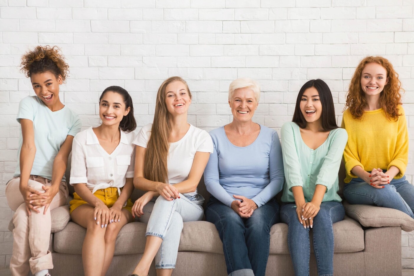 Six women of varying ages are sitting on a couch in front of a white brick wall, laughing and smiling, creating a joyful and lively atmosphere. They are dressed in casual attire with a mix of pastel and bright colors, as if ready for an outing to the best hair salon Cary NC boasts.