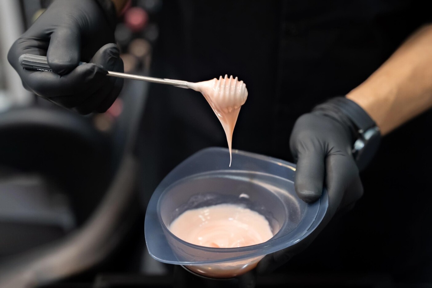 A colorist expertly wearing black gloves holds a brush coated in pastel pink hair dye, poised above a small bowl brimming with the vibrant hue.