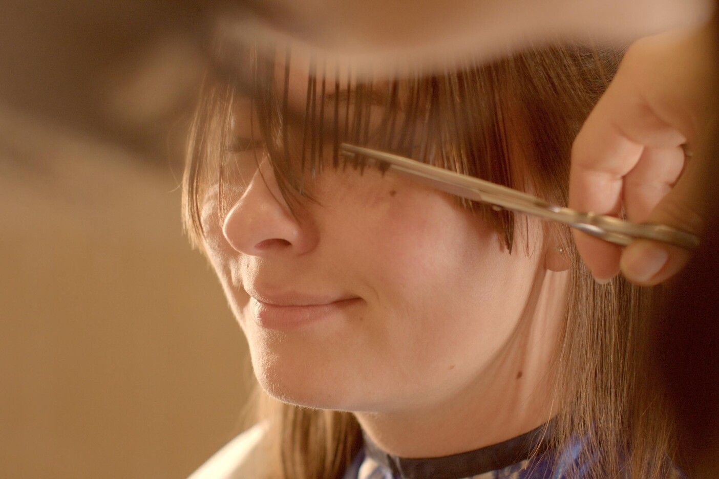A person with long brown hair is getting a precision haircut. Scissors are expertly trimming their bangs while they sit, smiling slightly, covered in a blue and white cape. Their eyes are obscured by hair being cut—a perfect snapshot of modern lifestyles embracing stylish solutions.