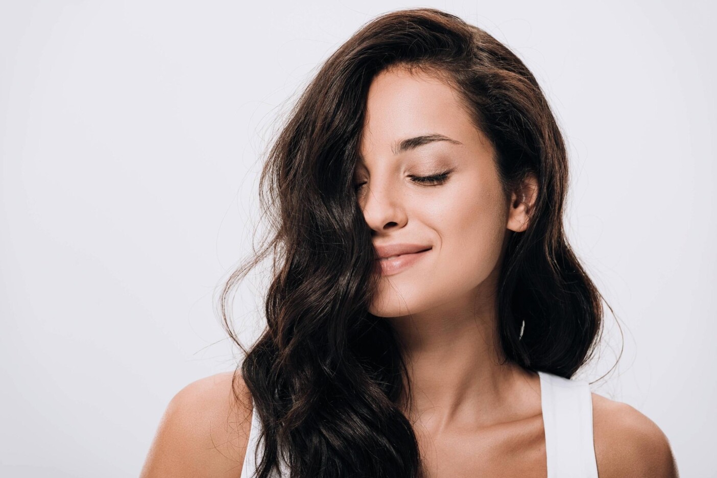 brunettes A woman with long, wavy dark hair and a slight smile poses with her eyes closed against a plain light background. She is wearing a white top.