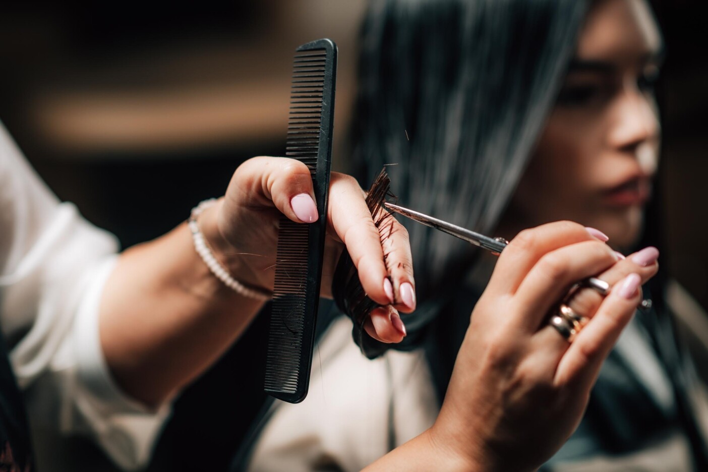 Beautiful Young Woman in Hair Salon A close-up of a hairdresser’s hands expertly delivering precision haircuts, cutting a client's dark hair with scissors and a comb, carefully sectioning each strand between their fingers. The client is blurred in the background.