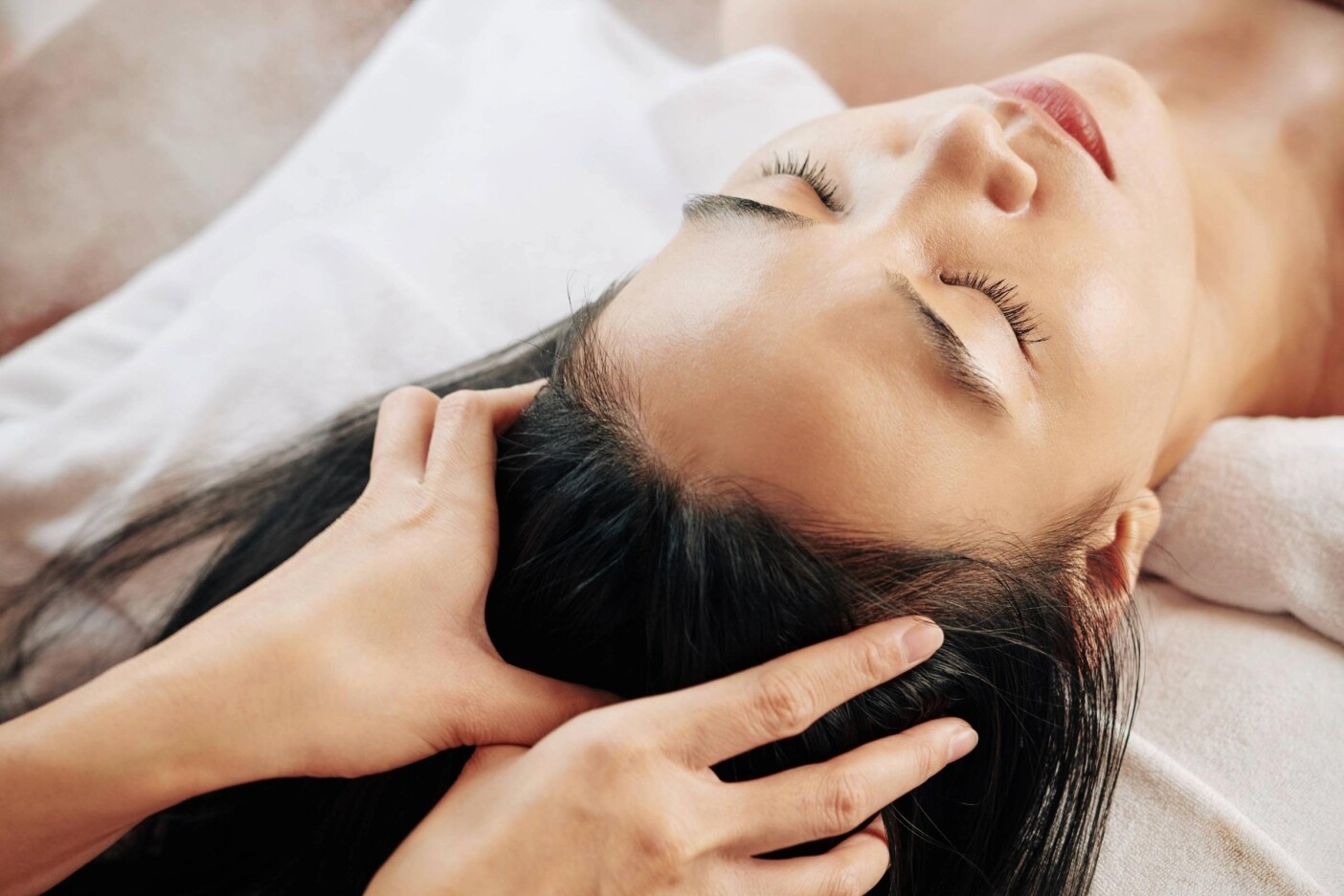 scalp.massage A person with closed eyes receives a relaxing scalp massage while lying on a white towel, conveying a sense of calm and wellness.