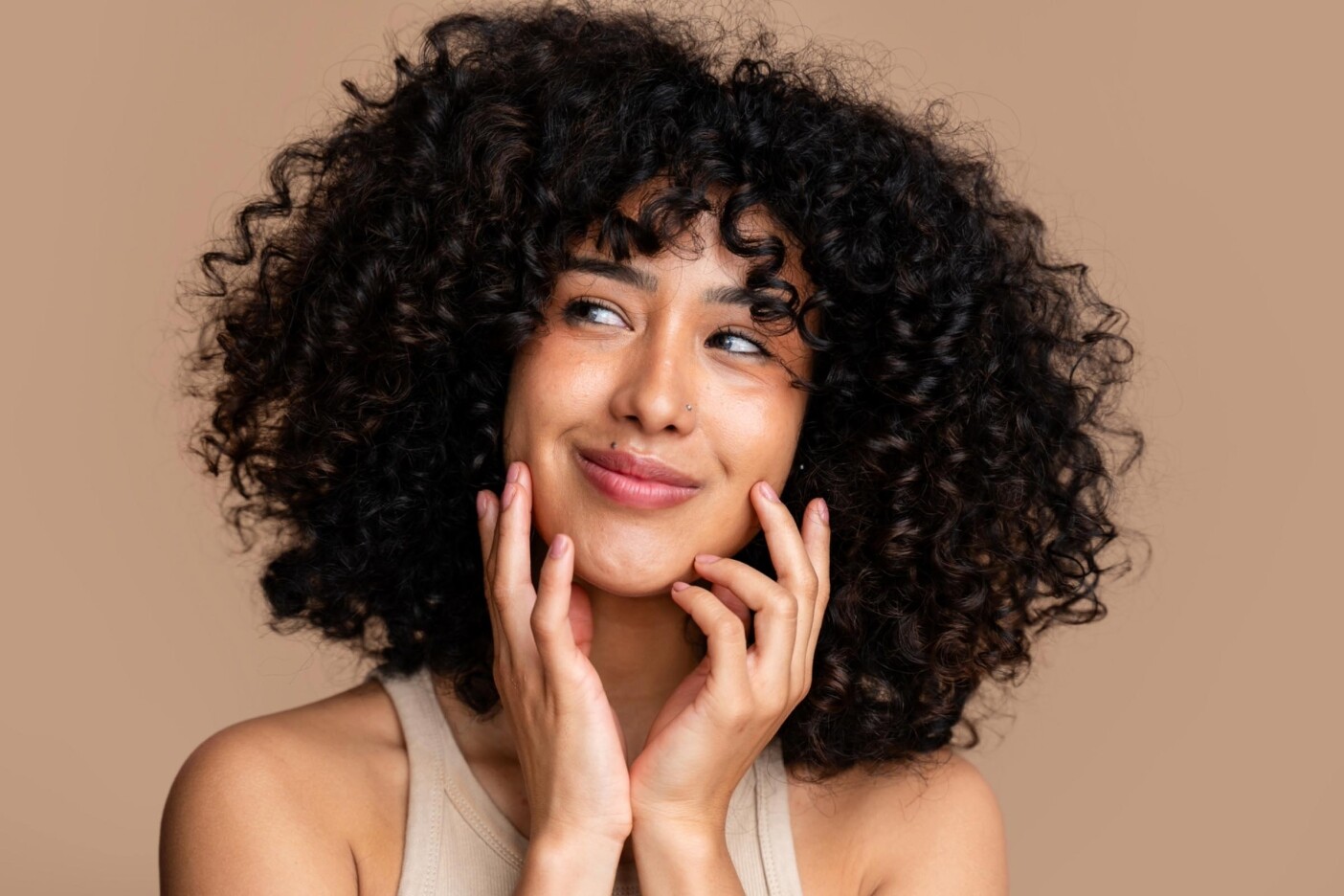 A young woman with curly hair near me smiles softly and looks to the side, gently touching her face with both hands. She is wearing a sleeveless top against a plain beige background.