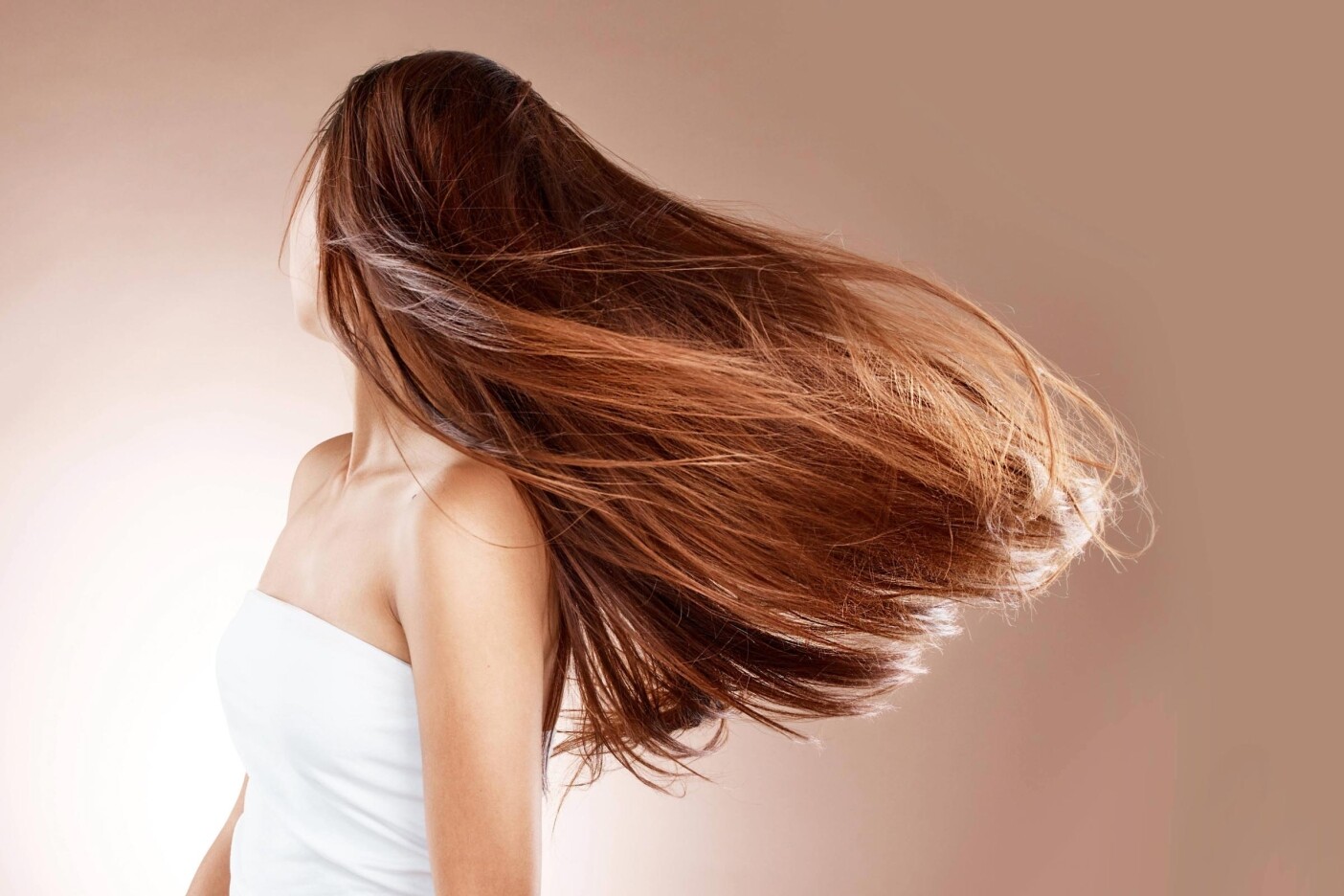 A woman with long, straight, brown hair flips her head, causing her hair to fan out. She is wearing a white, strapless top and is set against a soft beige background, capturing the effortless style from a Cary hair salon. Her face is turned away from the camera.
