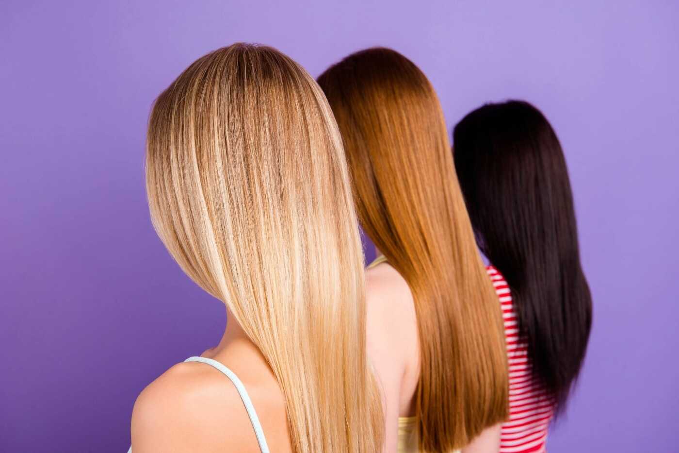 Three women with long, straight hair are shown from behind against a purple background at best hair salon Cary NC. One has blonde hair, another has red hair, and the third has black hair. They stand closely next to each other in a slight diagonal line.