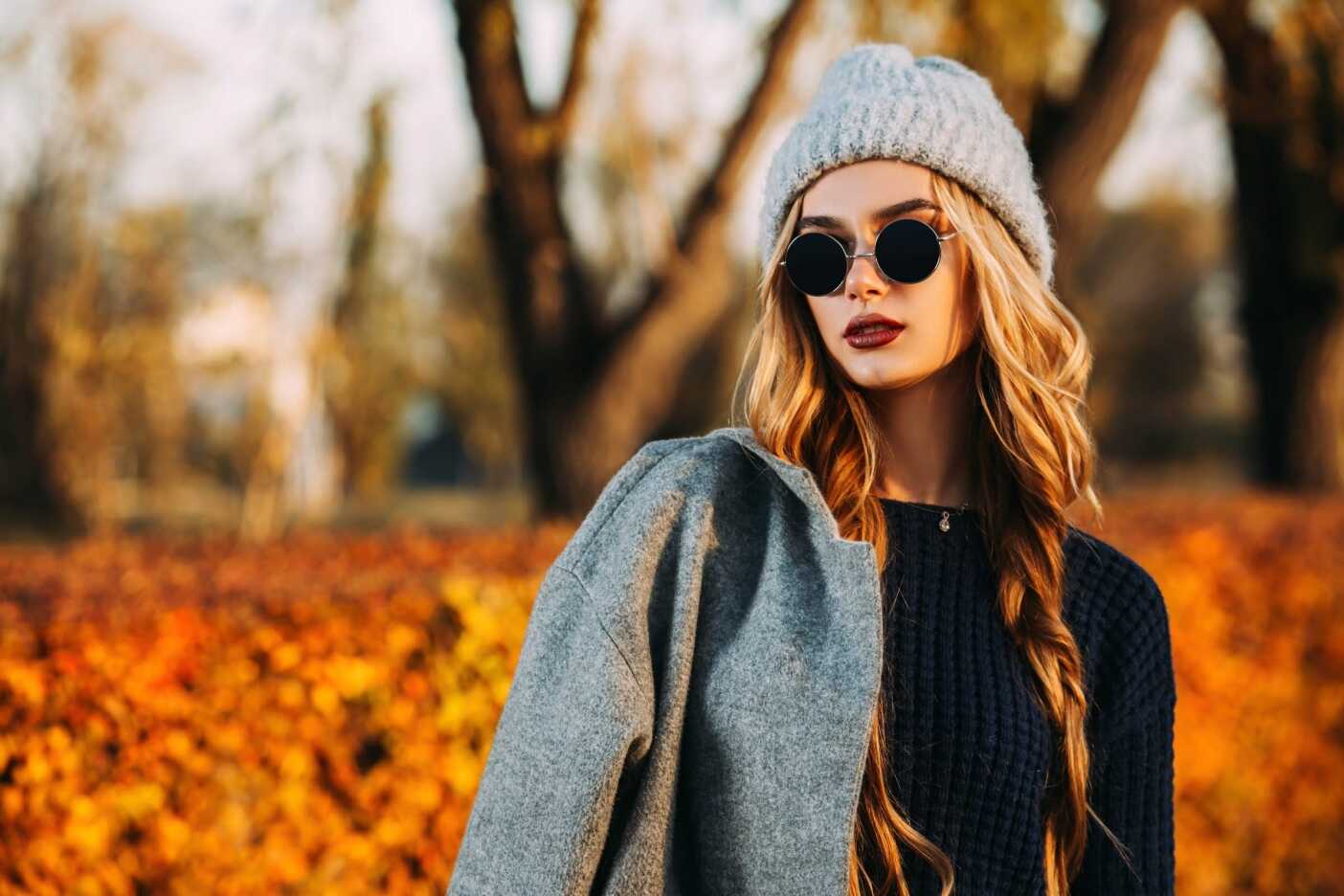 A woman with long blonde hair and wearing a white beanie and sunglasses stands outdoors. She is dressed in a dark sweater and has a grey coat draped over her shoulder. The background features autumn foliage and trees, showcasing the perfect look you'd achieve after a visit to the best hair salon Cary NC offers.