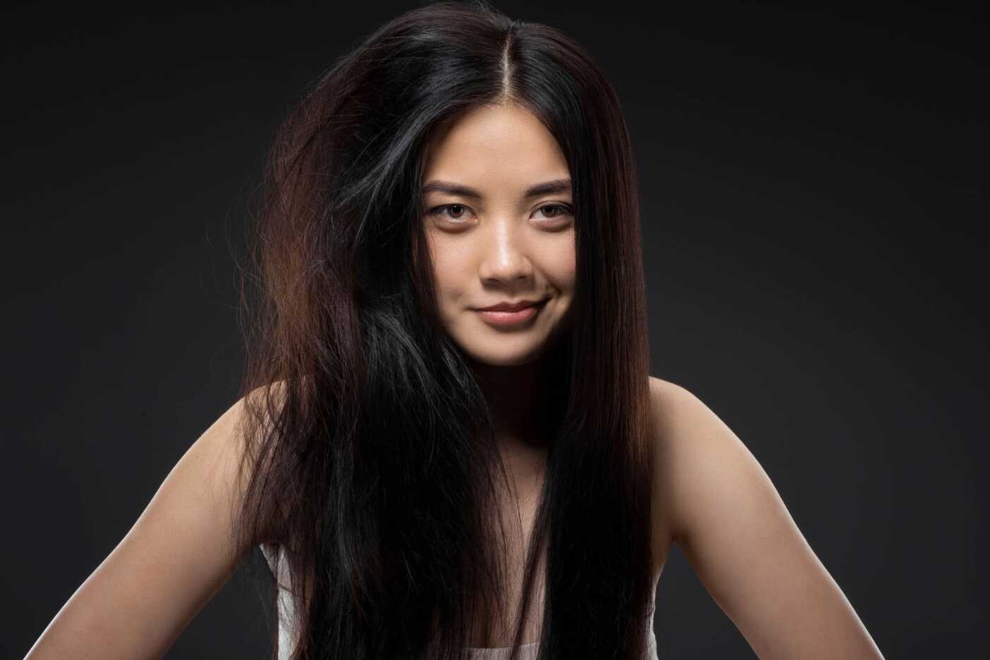 A woman with long, dark hair stands against a dark background. One side of her hair is smooth and shiny, while the other side is frizzy and unkempt, highlighting a distinct contrast. She is wearing a white top and looking directly at the camera with a slight smile, showcasing why hair salons in Cary NC are essential.