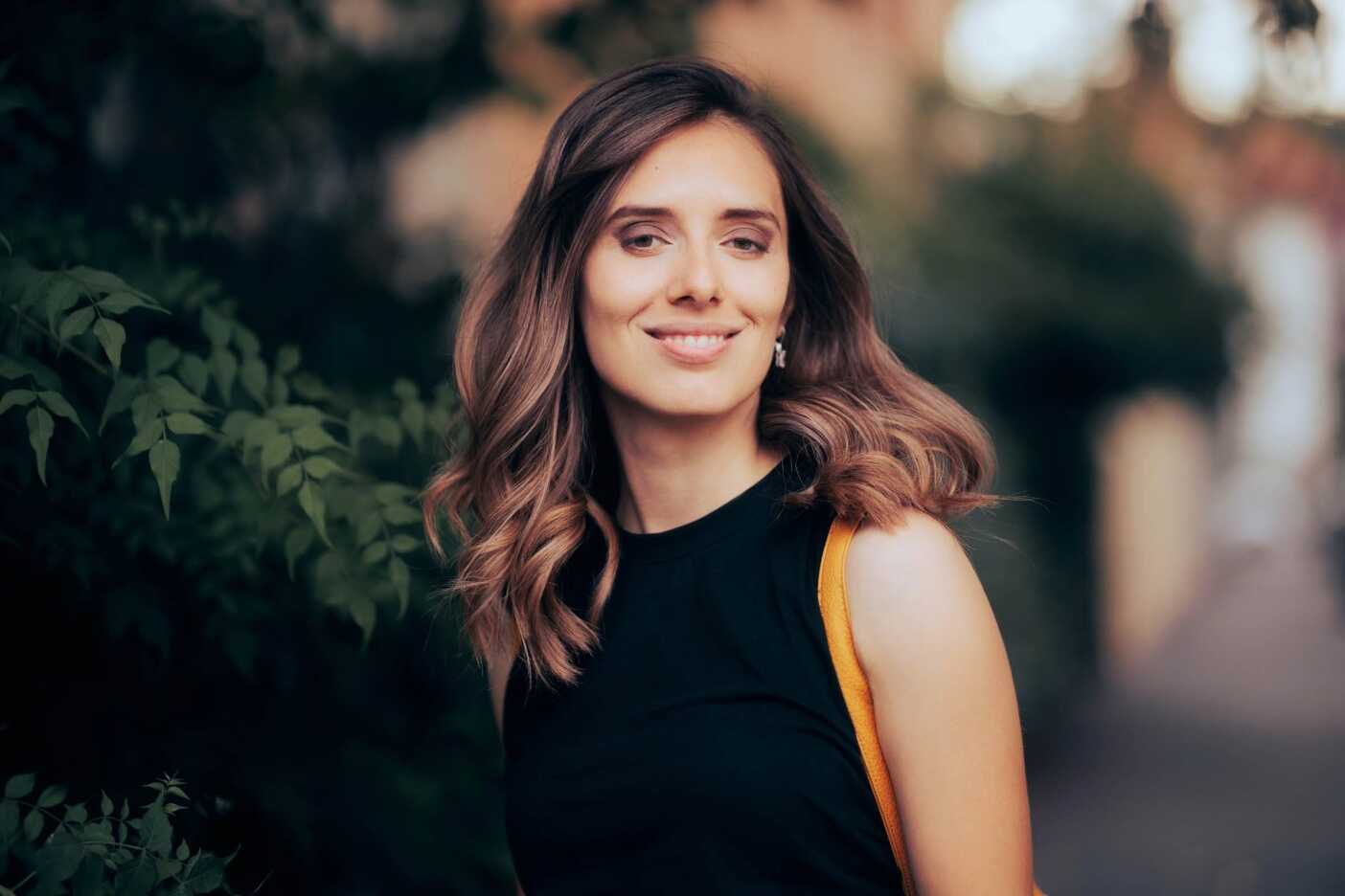 A woman with long, wavy, brown hair is standing outdoors, smiling at the camera. She is wearing a sleeveless black top and a yellow bag is visible on her shoulder. The background is blurred greenery and a path, possibly near one of the lovely hair salons Cary NC offers.