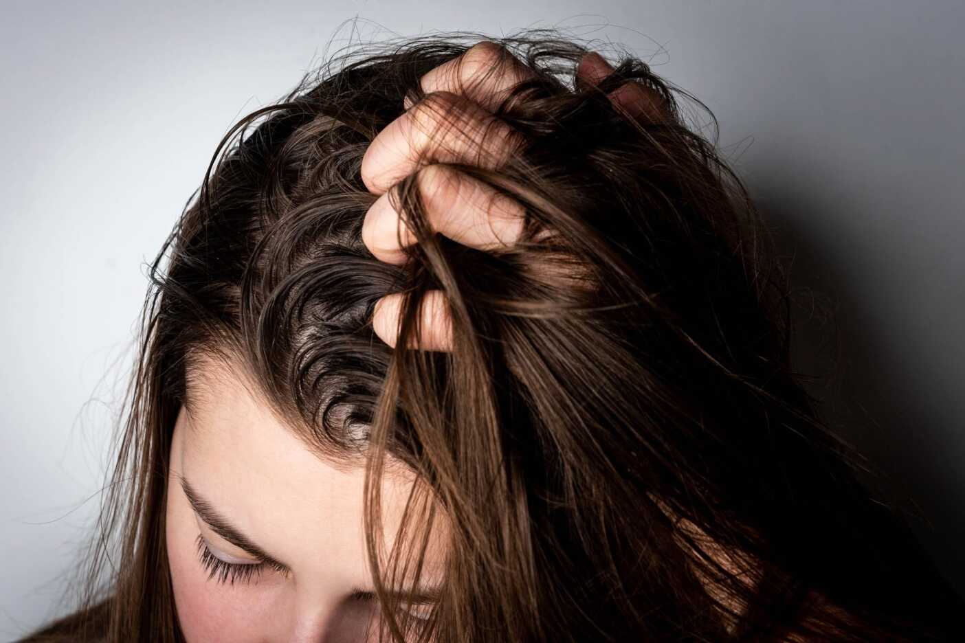 Close-up of a person with long brown hair pulling at their hair with one hand, their head bowed down and face partially covered by hair, perhaps in need of advice from a nearby hair salon. The image conveys distress or frustration. The background is plain and softly lit.