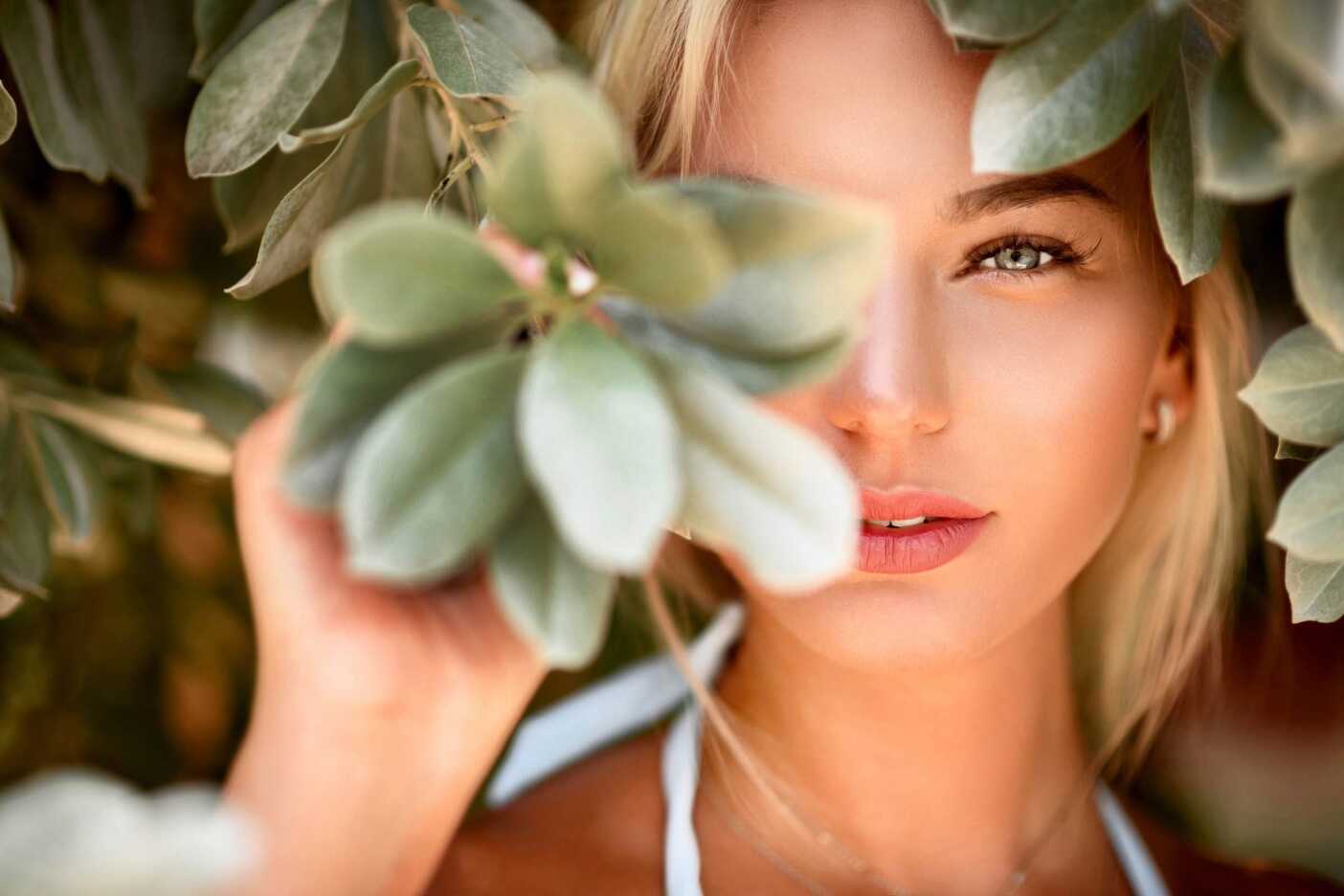 A close-up of a person with blonde hair partially obscured by green leaves. The individual's blue eyes and pink lips are visible, and they appear to be looking through the foliage. The background is blurred, emphasizing the person's face and the leaves in the foreground—perfect inspiration from a Sunday salon session.