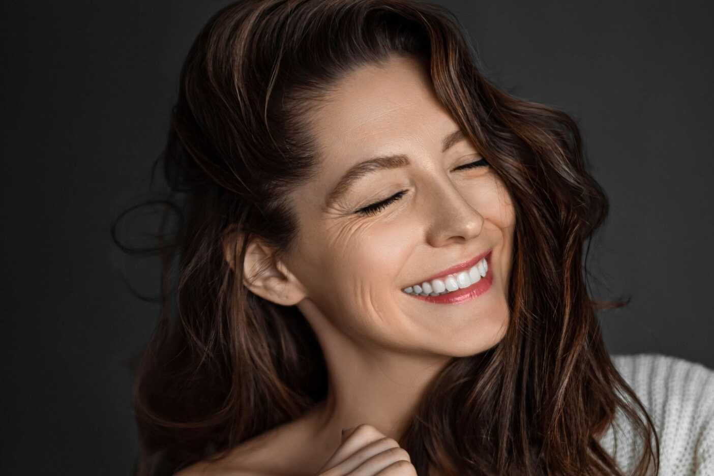 A woman with long, wavy brown hair is smiling with her eyes closed. She appears joyful and relaxed, as if she just stepped out of the best hair salon Cary NC has to offer, wearing a light-colored, off-the-shoulder top against a dark background.