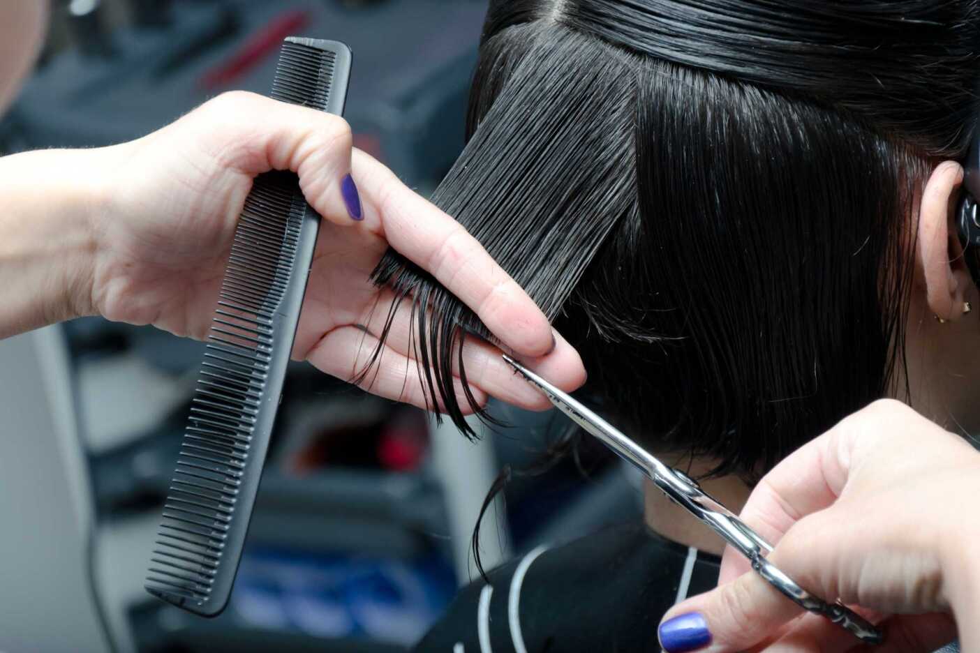 A close-up of someone with wet hair receiving a haircut in a cozy hair salon. One hand is holding a section of hair with a comb, while the other hand expertly maneuvers scissors. The background is slightly blurred and the person getting the haircut is wearing a black cape.