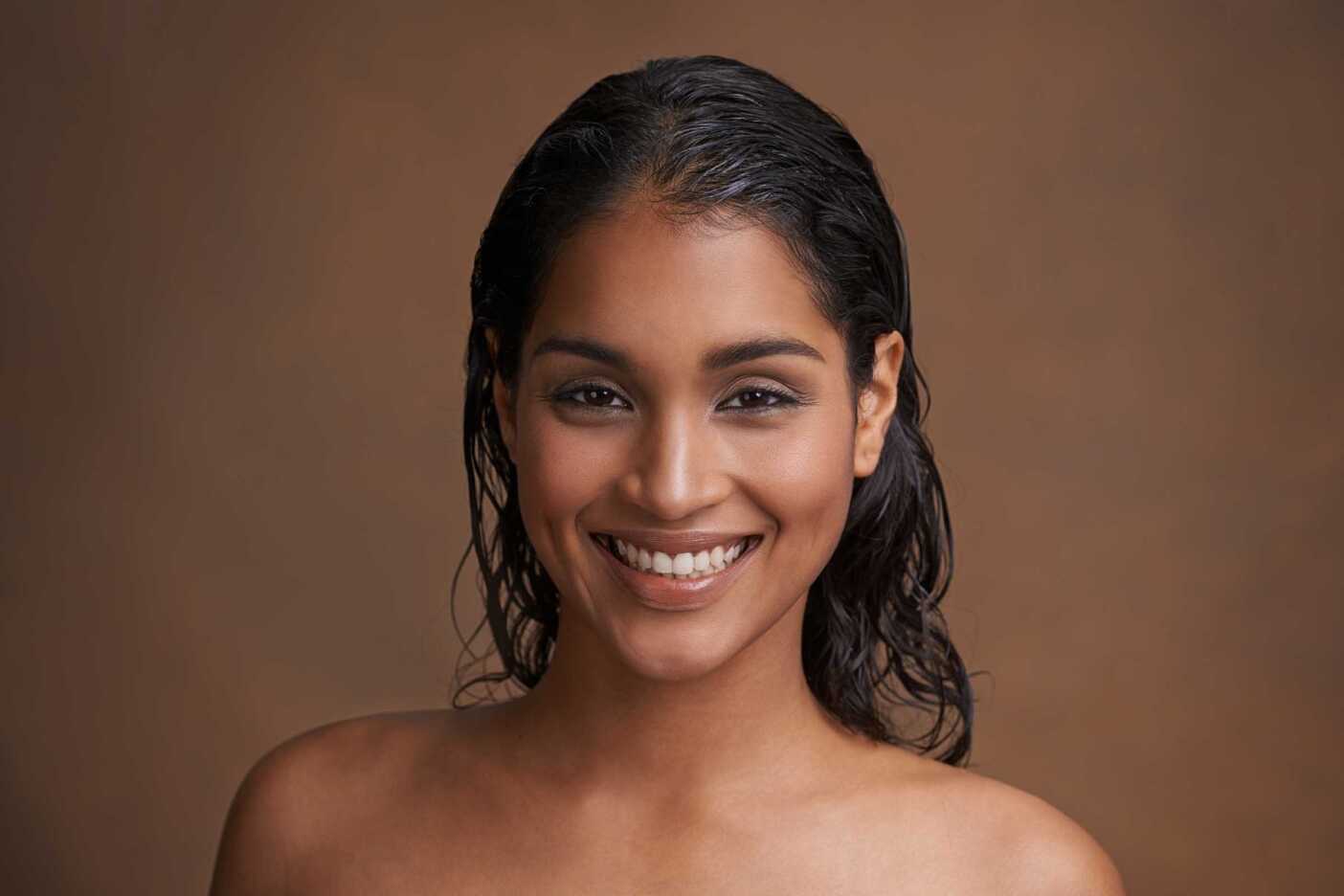 A person with wet dark hair and smooth skin smiling directly at the camera, possibly after a hair detox treatment at a hair salon in Cary, NC. The background is a warm brown color, and the person is not wearing any visible clothing. The image has soft, natural lighting.