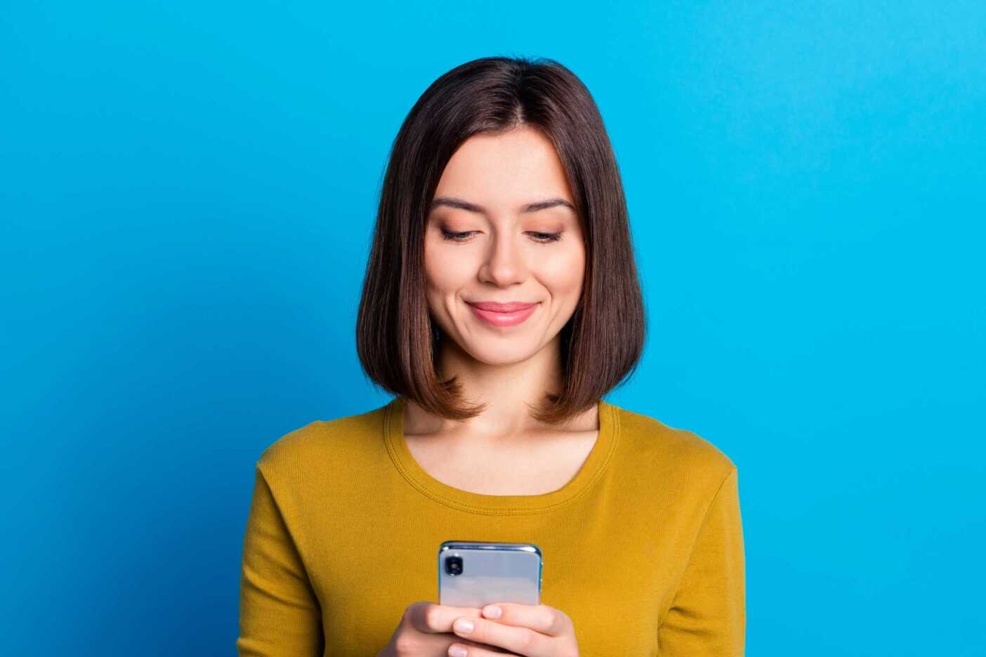 A woman with shoulder-length brown hair, wearing a yellow sweater, looks down at her smartphone and smiles, possibly delighted by booking her salon appointments online. The background is solid blue.