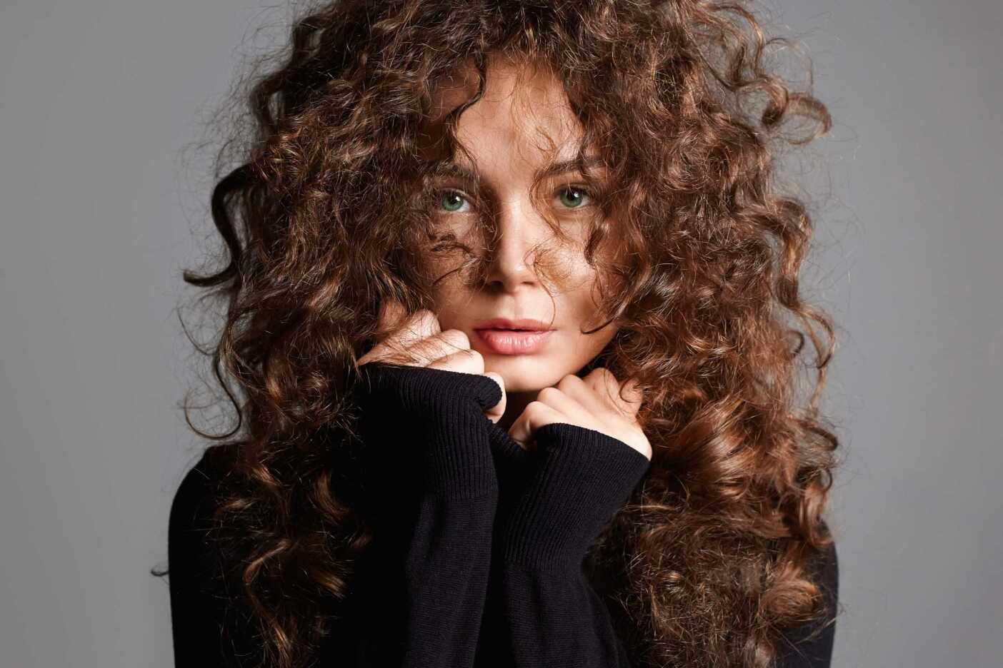 A person with the best curls, framed perfectly around their face, is wearing a black sweater and looking directly at the camera against a gray background. They hold the sweater up to their chin with both hands, showcasing impeccable hair care.