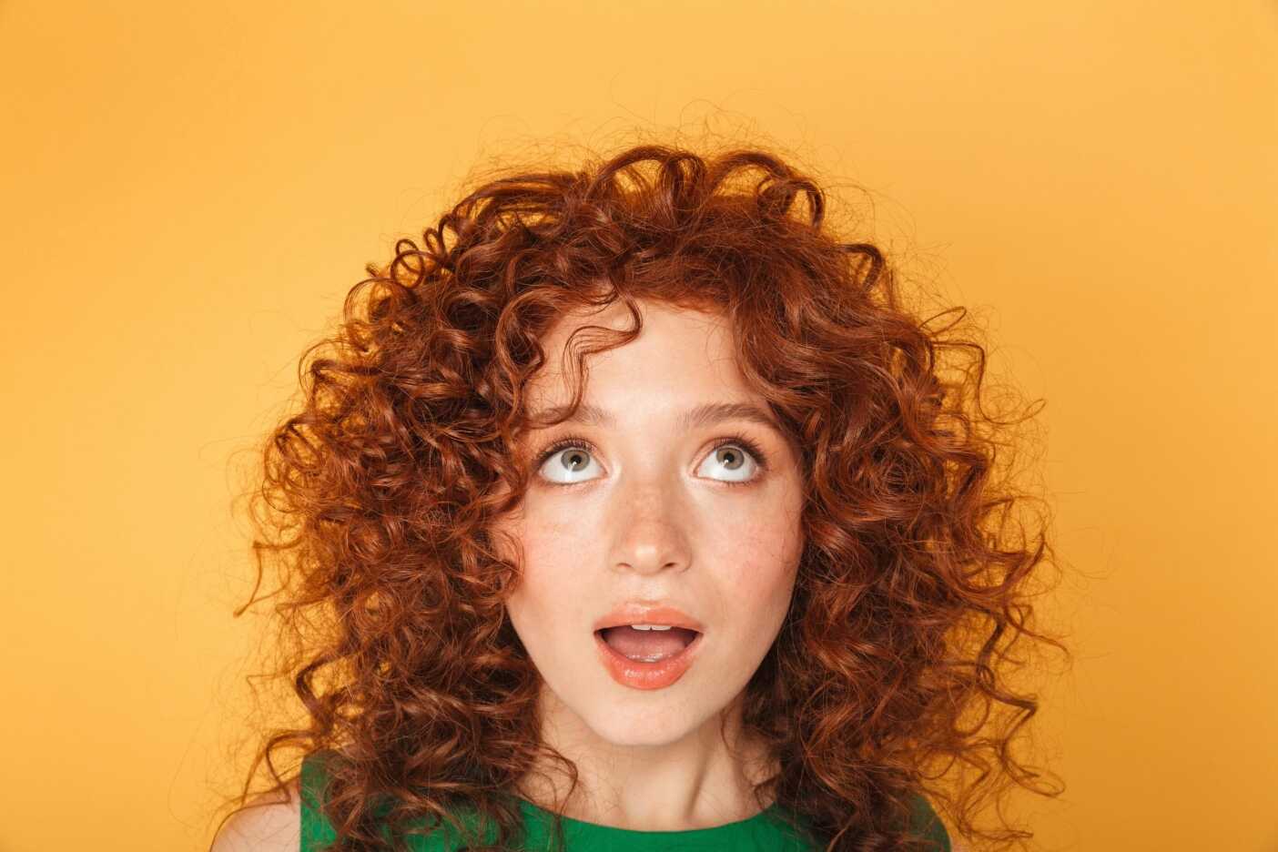 A person with a trendy curly haircut and red locks looks upward with a surprised expression, mouth slightly open. The vibrant background is a solid yellow, highlighting their green top perfectly.